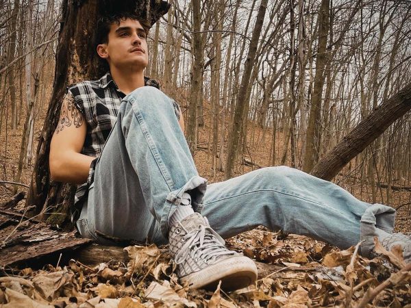 man sits against a tree in the forest wearing a plaid cutoff shirt and faded blue jeans