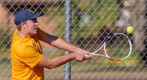 a player dressed in a gold t-shirt and blue hat hits a tennis ball