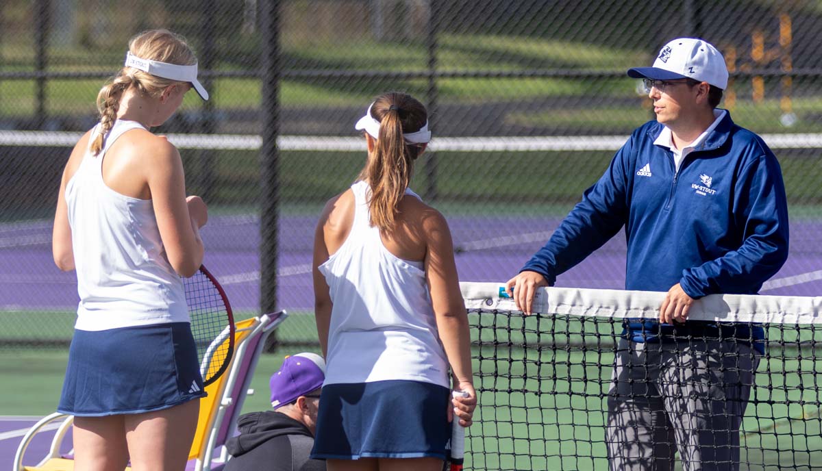 A tennis coach standing at the net talks to his doubles team between sets.
