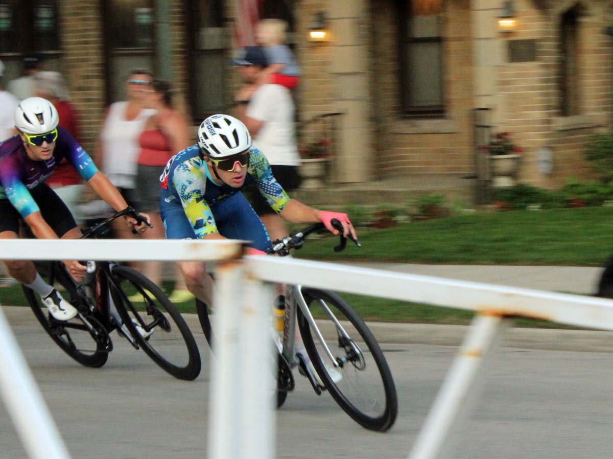 Two multi-color clad road-racing cyclists go around a corner in a small town.