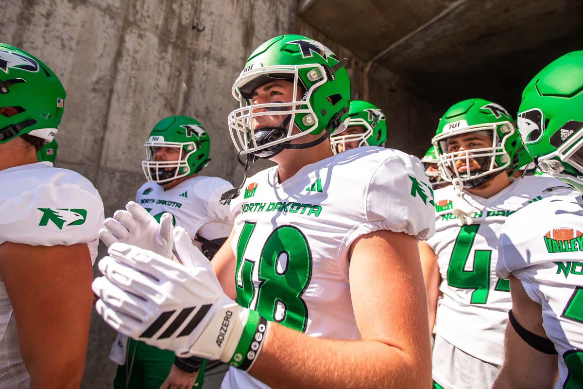 University of North Dakota football players prepare to take the field. The players wear white uniforms with green numbers and green helmets.
