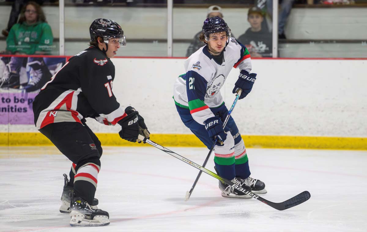 A men's hockey player in a dark uniform and a men's hockey player in a white uniform skate during a recent game.