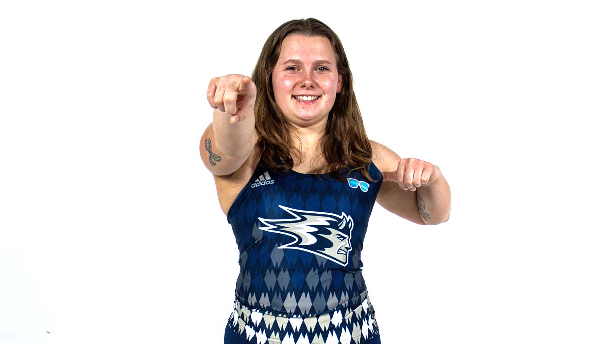 In a studio photograph, a member of the UW-Stout women's track and field team, wearing a dark blue uniform, points at the camera.