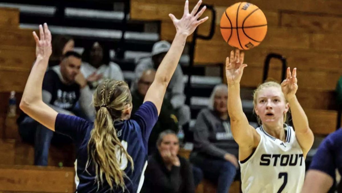 A women's basketball player in a white uniform takes a shot over an opponent in a blue uniform.