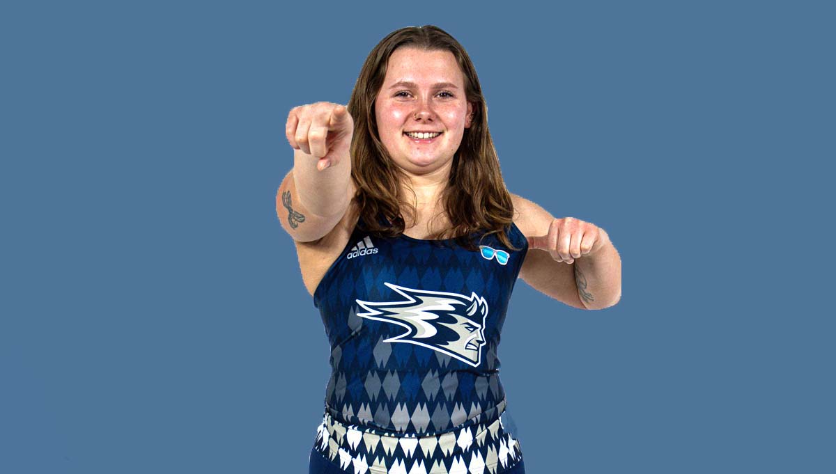 In a studio photograph, a member of the UW-Stout women's track and field team, wearing a dark blue uniform against a lighter blue background, points at the camera.