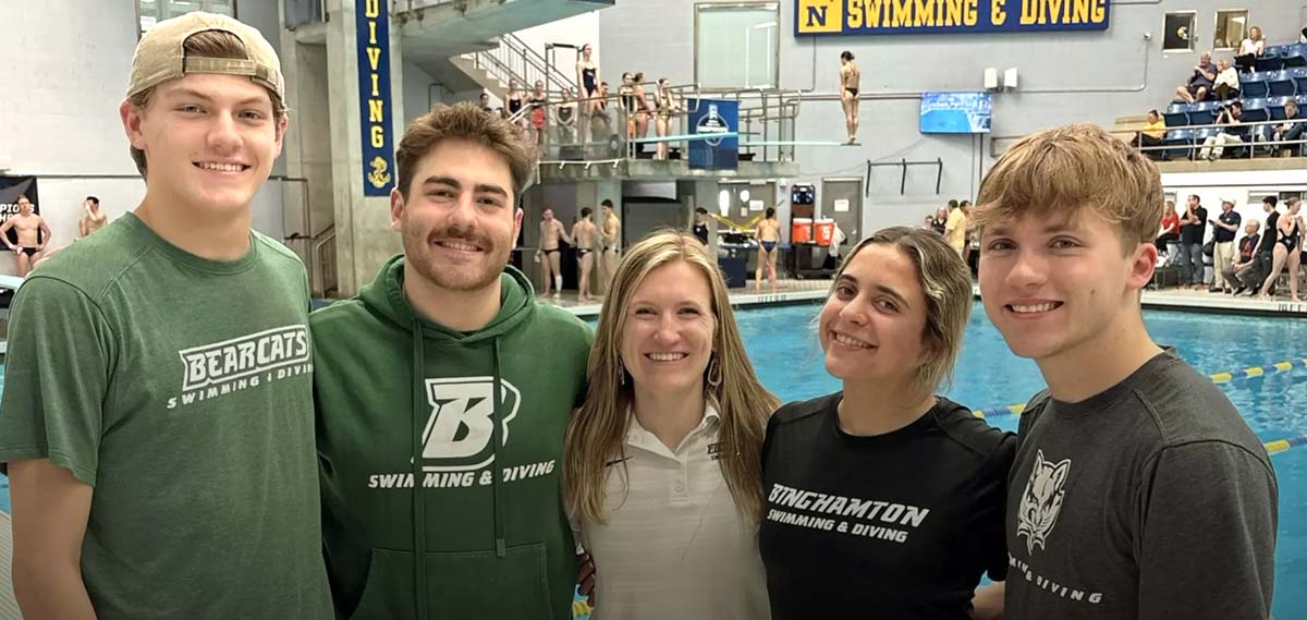 Five people, wearing green or white shirts, pose for a group photo while diving competition continues in the background.