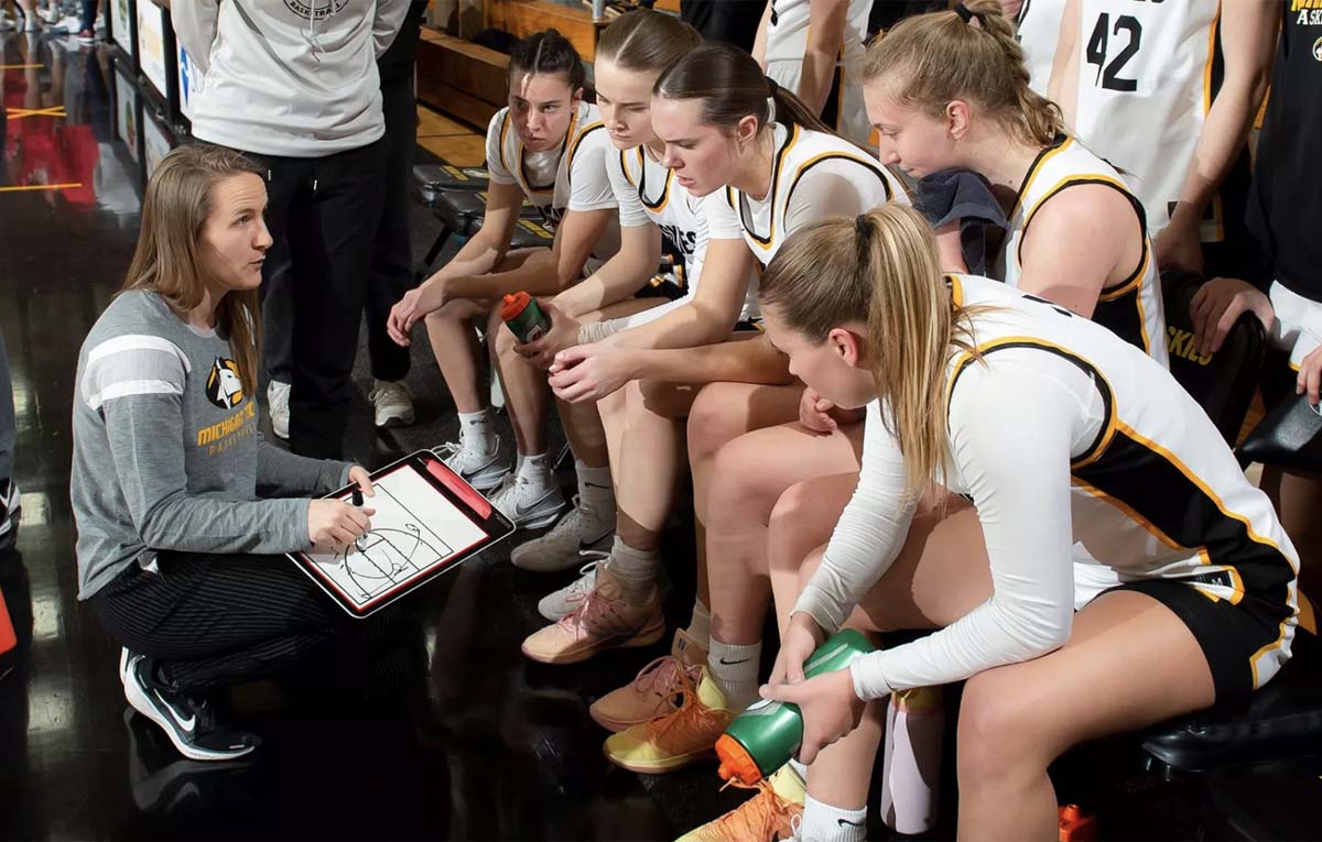 A kneeling women's basketball coach explains a play to her team during a timeout.