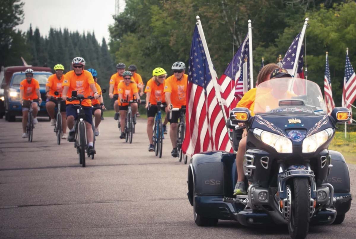 Dressed in orange, bicycle riders Highground Heroes Ride follow a lead motorcycle draped in US flags.